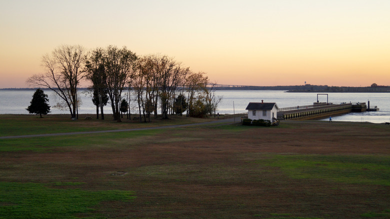 Shoreline of Fort Mott State Park at sunset, with a white building beside a pier, sparse trees to the left, and brown and green grass in the foreground