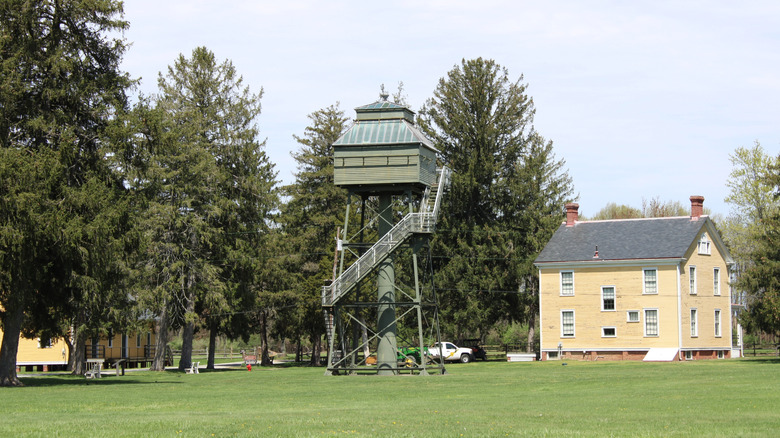 Green fire tower and yellow house in Fort Mott State Park, surrounded by grass with trees and a white truck in the background, under a pale blue sky