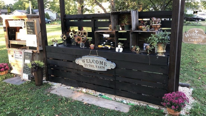 Wooden farm stand with flowers in vases and squash in a box, with a sign saying "Welcome to the Farm", surrounded by grass with homes, cars, and trees in the background