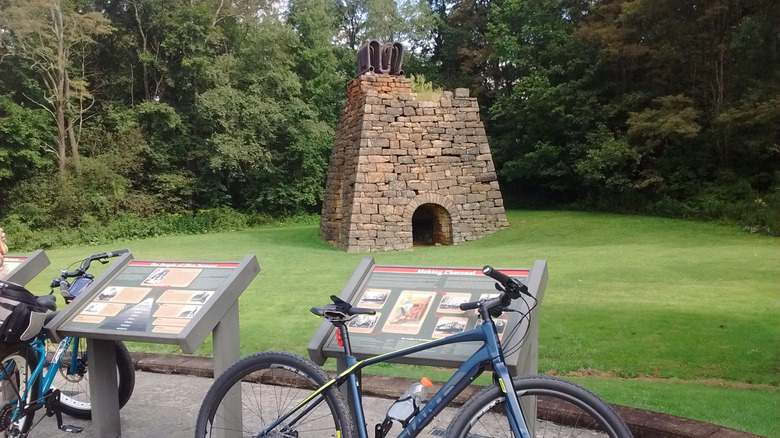 Eliza Furnace, a trapezoidal stone structure with metal pipes on the top, surrounded by grass with trees in the background and interpretive signs in the foreground with blue bikes leaning on them