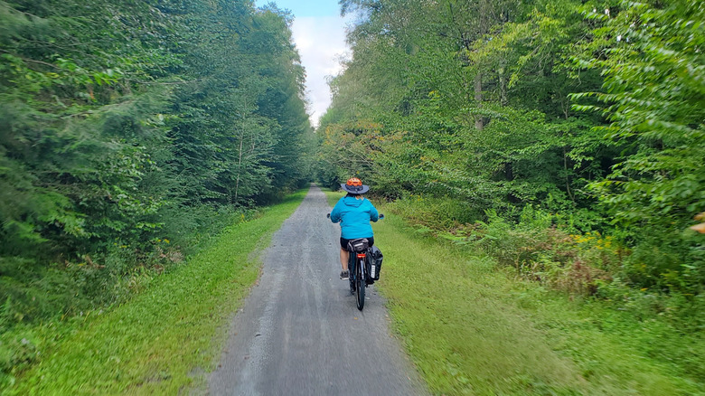 Person in a blue jacket and wide-brimmed black and orange hat rides a bike down the Ghost Town Trail, with trees and grass growing to either side