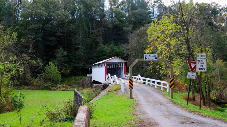 Jackson MIll Covered Bridge, a white-painted covered bridge on a dirt road, with clearance and weight limit signs in the foreground, green grass around it, and trees in the background