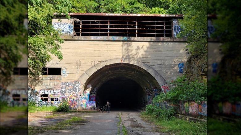 Biker in a brown shirt prepares to enter Sideling Hill Tunnel, a gray stone structure with graffiti surrounded by trees