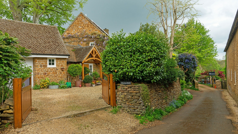 A narrow country lane in Hook Norton, Oxfordshire, UK, shows a stone cottage behind a well-manicured stone wall.