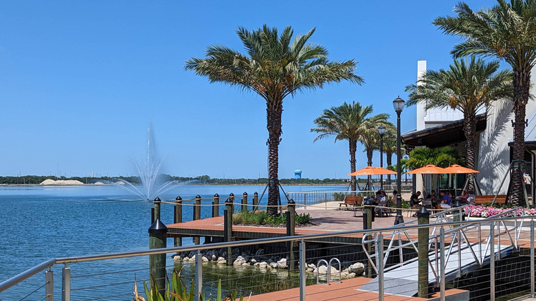Outdoor dining near Waterside Park in Lakewood Ranch, Florida, beside blue water and a luxurious fountain with blue sky and date palm trees