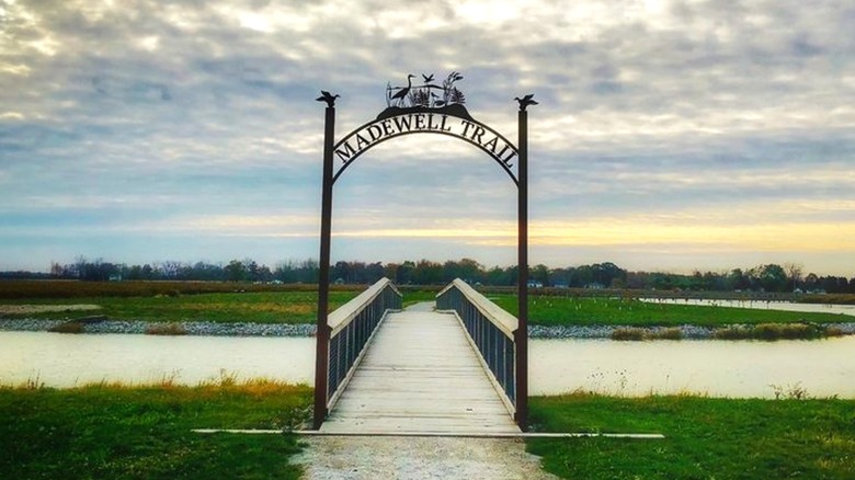 An arch sign that labels the Madewell Trail in Howard Marsh over a wooden bridge going over water and leading to a grassy area.