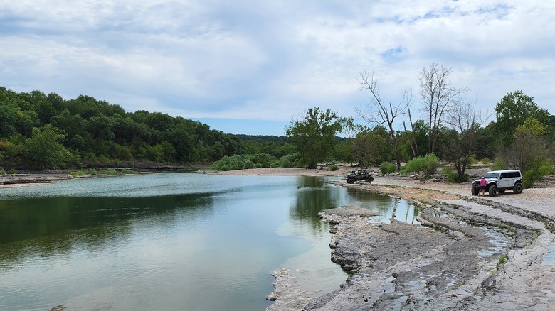 An off-road vehicle and a jeep sitting on the rocky shore next to a wide shallow river with green trees on the opposite bank