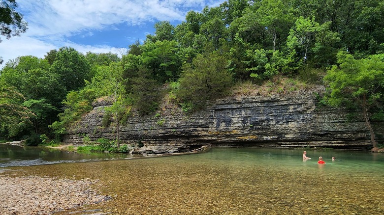 Three young people sitting in the clear water of a river at the Little Blue State Park Area with a rocky, tree-topped wall behind them