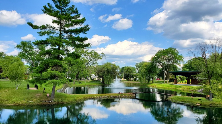 Pond in Pauquette Park, Portage, with a fountain and lined with vibrant green grass, trees, and a pavilion