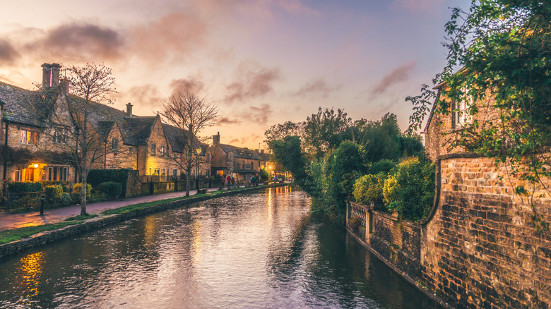 A canal running through Bourton-on-the-Water, with lit up homes flanking it on the left, under a sunset colorful sky