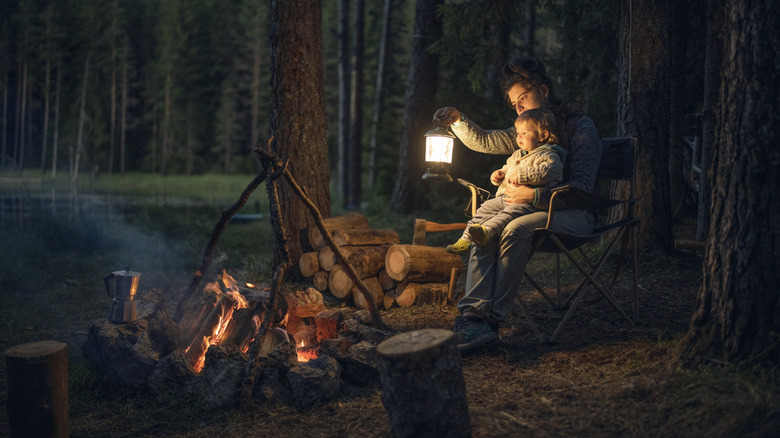 Woman holding camp lantern with her child on her lap by a campfire