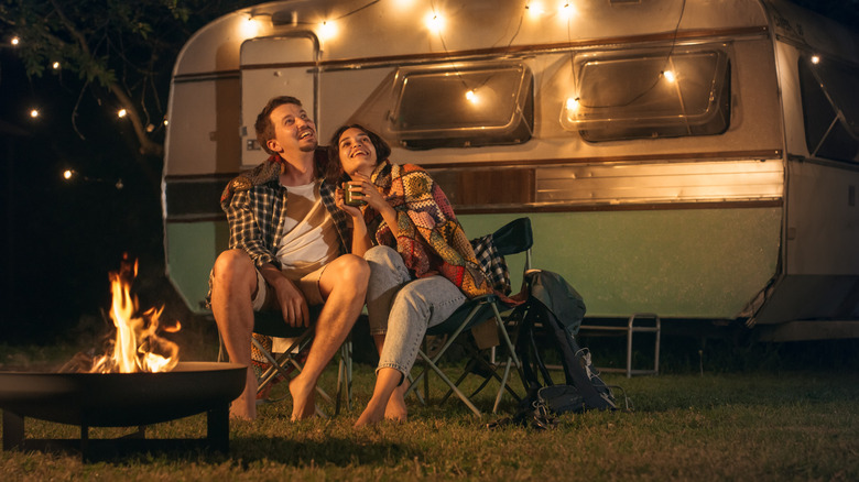 A smiling couple sits by a campfire and trailer with string lights.