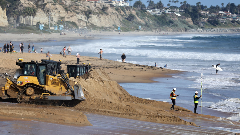 Workers using bulldozers to move fresh sand into place onto a California beach with beachgoers in the background.