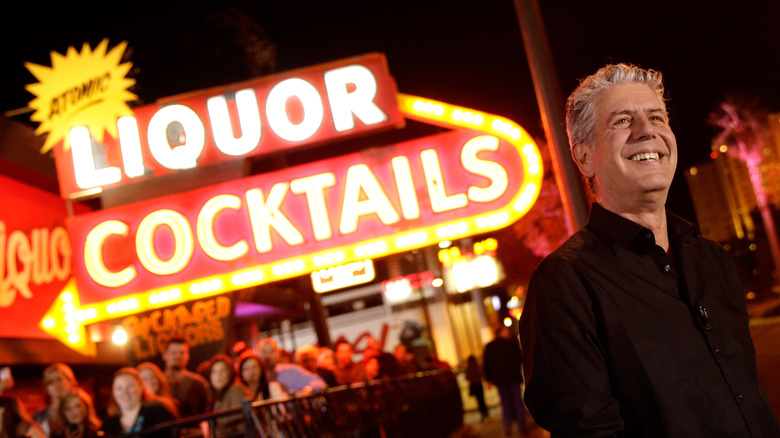 A smiling Anthony Bourdain in front of a big neon sign that says liquor and cocktails with a big yellow arrow