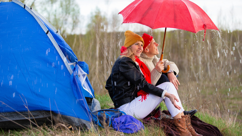 An elderly couple under an umbrella next to their tent in the rain