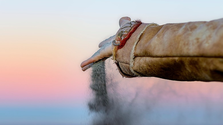 Hand of a person wearing a brown jacket scattering ashes into the air at sunset