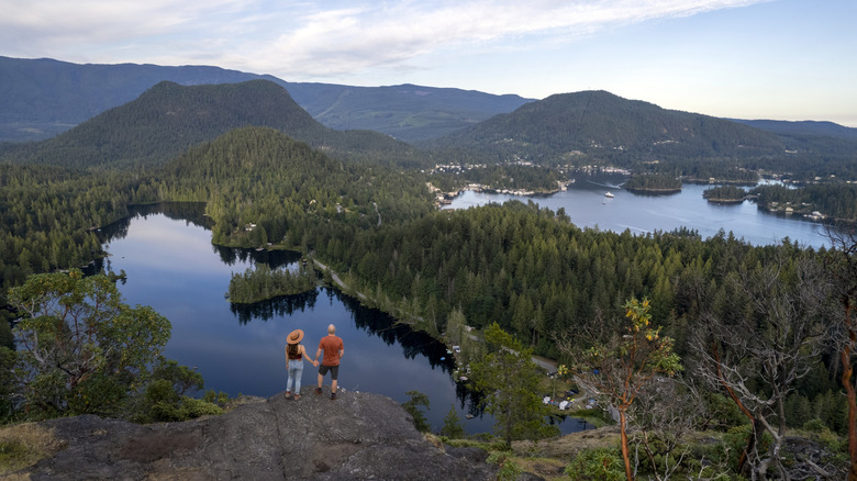 A couple stands on the end of a rocky ledge, looking out over a beautiful landscape of lakes, forests, and mountains