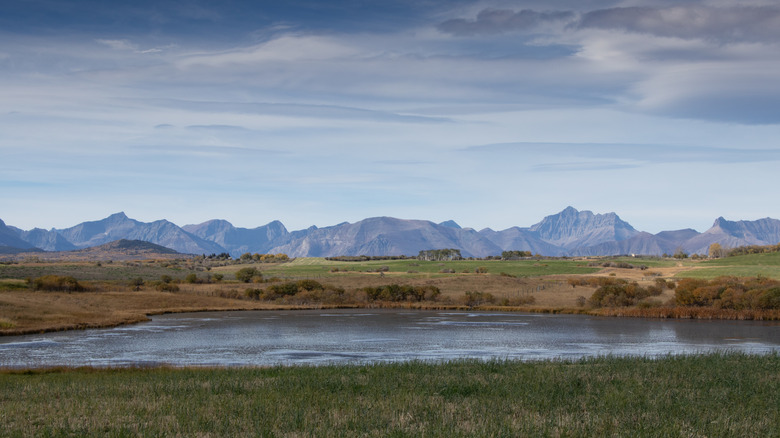 View of a rocky mountain range along the horizon, with a lake and green fields in the foreground