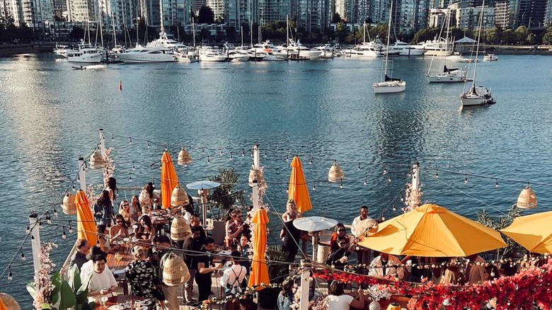 Patrons on a sunlit patio on the water with umbrellas, tables and heat lamps.