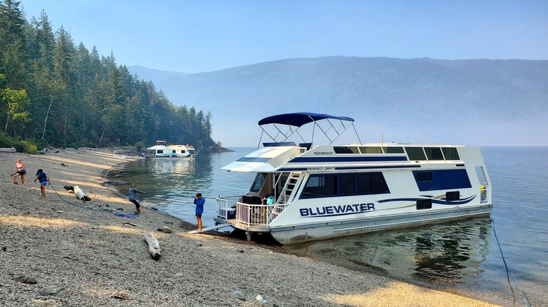 A two-level houseboat anchored at a rocky beach, with people walking on shore, pine trees behind, and the opposite shore in the distance