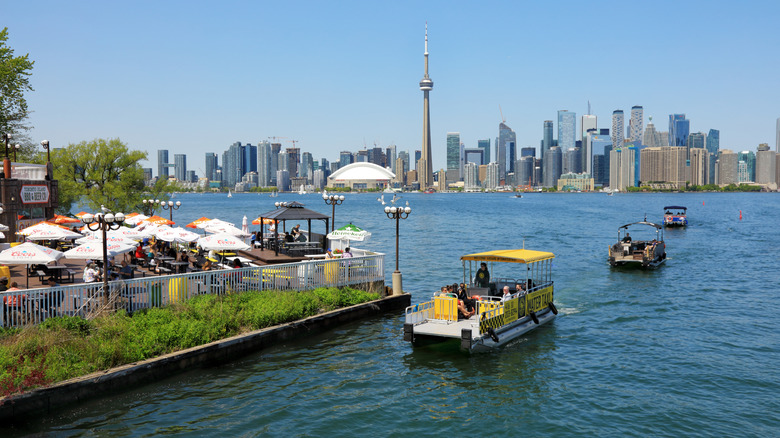 A sunny patio at Toronto Island BBQ & Beer Co. with small water taxis on the harbor and views back to Toronto's city skyline and the CN Tower.