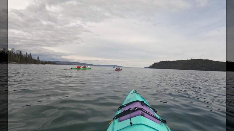 The front of a kayak with two kayaks in front and the expansive lake beyond them with tree-covered shores in the distance