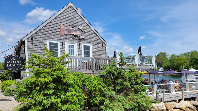 Side view of The Rope Loft restaurant, a building with outdoor seating and Cape Cod style shingle architecture, in Chester, Canada, with green trees and a vibrant blue sky with clouds