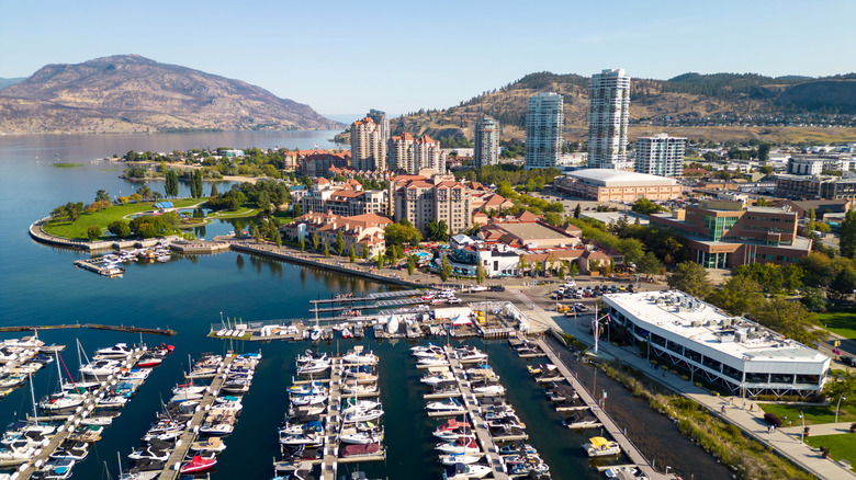 Aerial view of a lakeside marina with boats, high-rise buildings, and surrounding mountains in Kelowna, Okanagan Valley