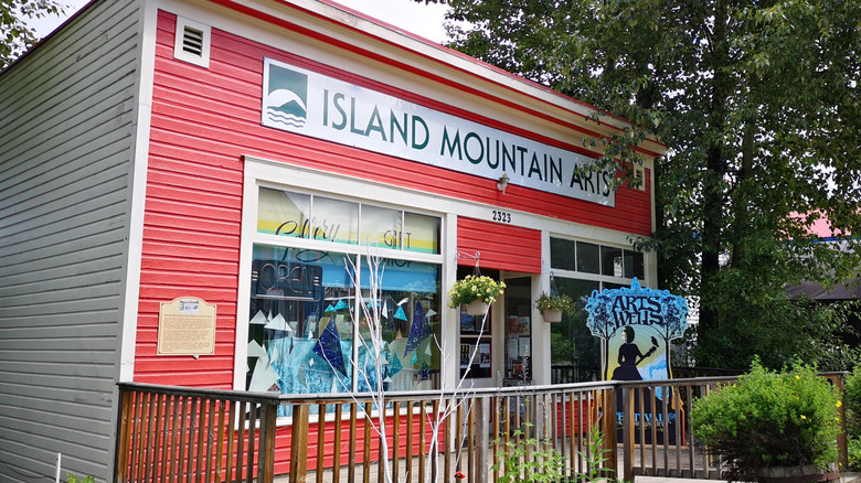 Red-painted facade of the Island Mountain Arts center in Wells, with art-filled windows and trees in the background.