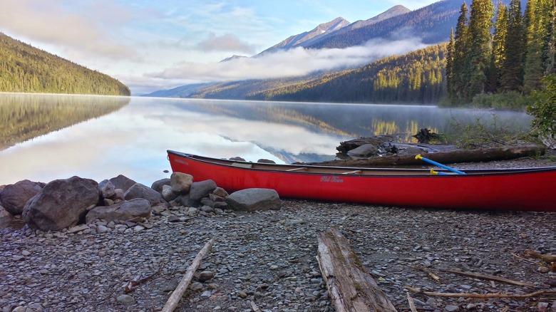 A red canoe sits on a rocky shore in front of a beautiful lake surrounded by green forests and mountains.