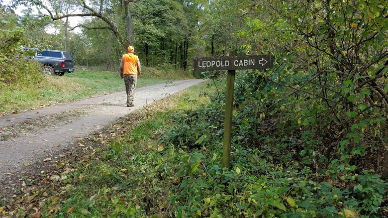 Man wearing hunting gear walking trail with sign to Leopold cabin in Missouri
