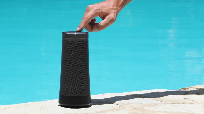 A hand presses play on a black Bluetooth speaker by a swimming pool.