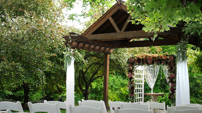 A flower-adorned wedding arch with plants and trees and grass around it.