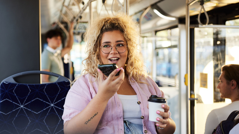 A person with glasses holding a coffee and chatting on speakerphone while taking public transit