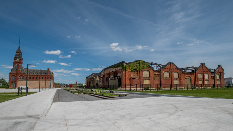 Abandoned Pullman Factory, a red brick building with a missing roof, boarded windows, and ivy, beside a smaller red brick building with a clock tower, surrounded by grass and sidewalks under a blue sky