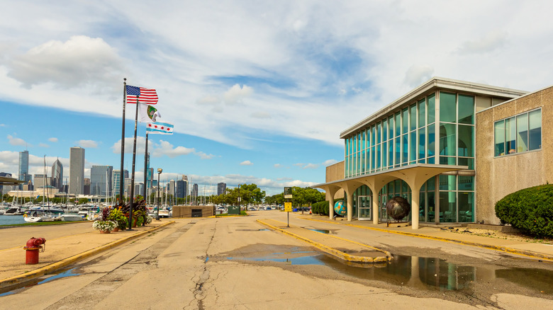 Meigs Field Terminal along the right, a tan building with large windows, with flagpoles and the Chicago skyline on the left, under a cloudy blue sky