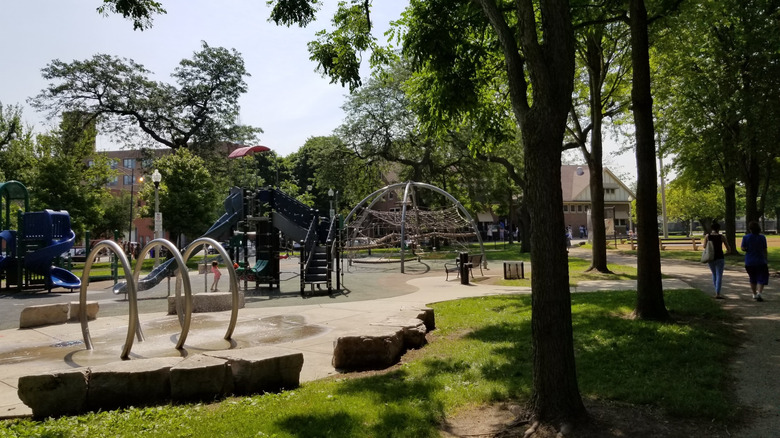 Playground in Union Park, with spray rings, slides, and a climbing net surrounded by grass and trees, with buildings in the background under a blue sky