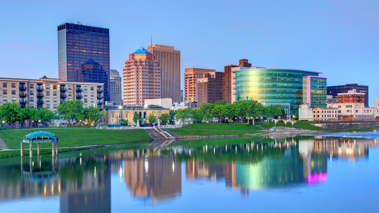 Downtown Dayton, with several tall buildings behind a green grassy area along the Miami River, under a blue sky