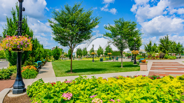 A park in Dayton, with green vines and a black lightpole with flower baskets in the foreground, a green lawn with trees surorunded by gray and red paving stones in the center, and white pavilion domes in the background, under a cloudy blue sky