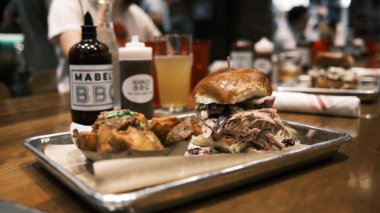 A tray on a table with a pulled pork sandwich and side of potatoes with a bottle of barbecue sauce and drinks in the background.