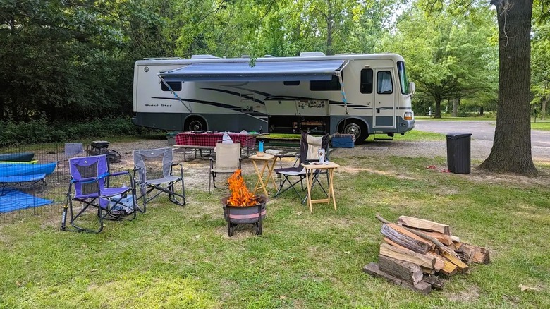 An RV surrounded by a fire and folding chairs, parked inside a wooded area at Clinton Lake in Illinois.