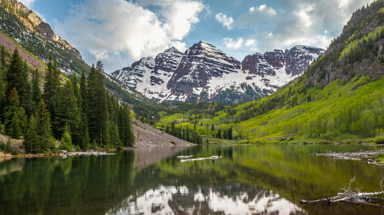 Majestic Maroon Bells rises above the serene Maroon Lake, surrounded by lush greenery and snow-capped peaks under a blue sky