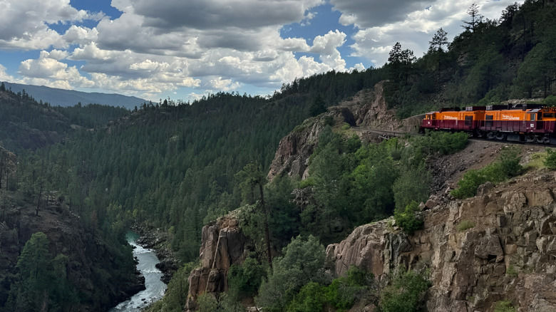 A train travels along a rocky mountain ledge above a river and forest valley near Needleton, Colorado