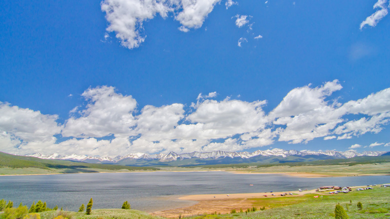 Cars and boats at Taylor Park Reservoir with surrounding snow-capped mountains and a blue sky.