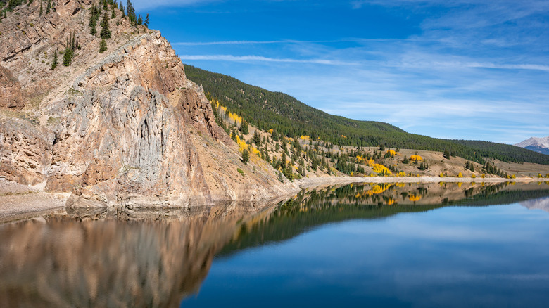 The calm water of Taylor Park Reservoir reflecting a rocky mountain and tree-covered hillside along its shoreline.