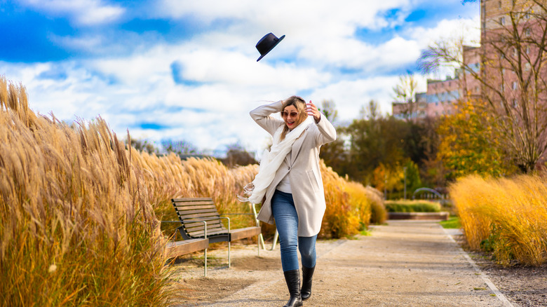 A woman walking in city park on sunny day in autumn smiling with surprise as the wind carries her hat into the air