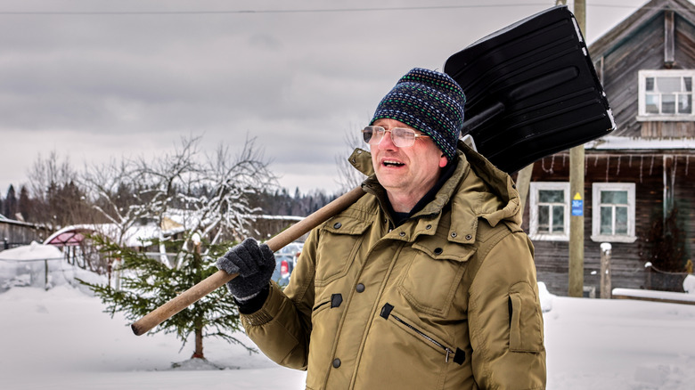 Man in olive green winter clothing holding a shovel with a snow covered landscape visible behind him