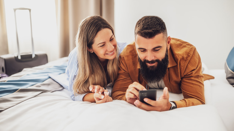 Young happy couple is on vacation. Tourists arrived in hotel room with their suitcases. They are lying on bed and using smartphon