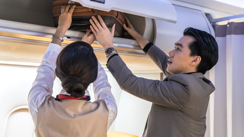 An air stewardess helping a man put his luggage in the overhead locker on a plane.