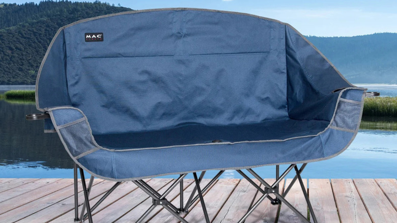 A blue Mac Sports Double Camping Chair sitting on a lakeside dock surrounded by nature.